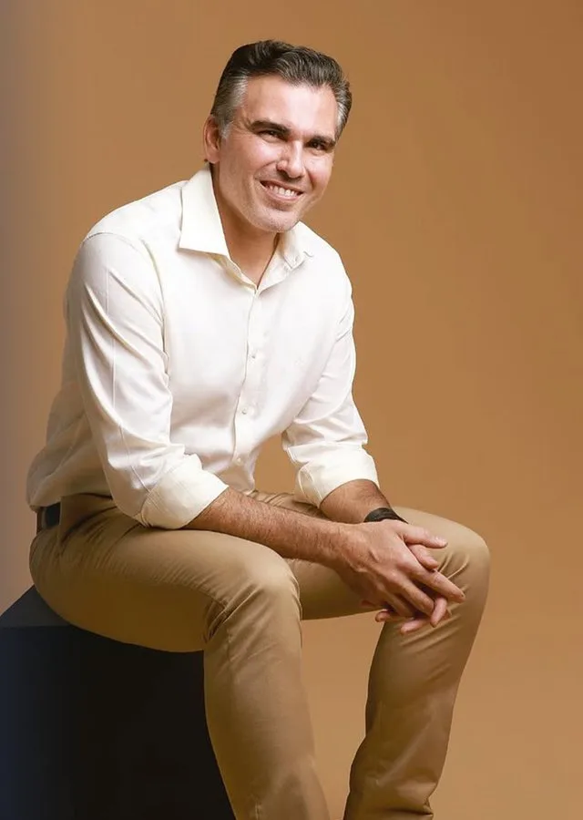 Dr. Fábio Saab sentado, de camisa branca e calça bege, em fundo marrom