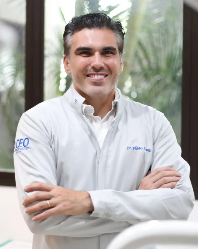 Dr. Fábio Saab de jaleco branco, sorrindo, com os braços cruzados