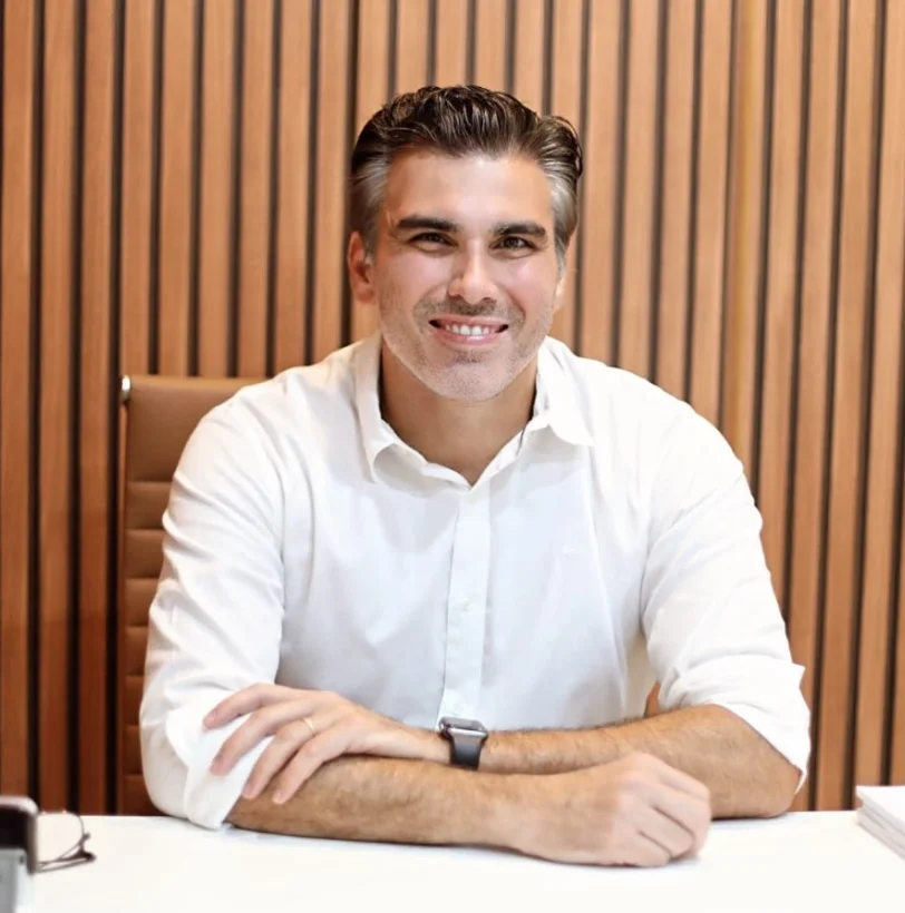 Dr. Fábio Saab de camisa branca, sorrindo, com os braços cruzados sobre a mesa do consultório